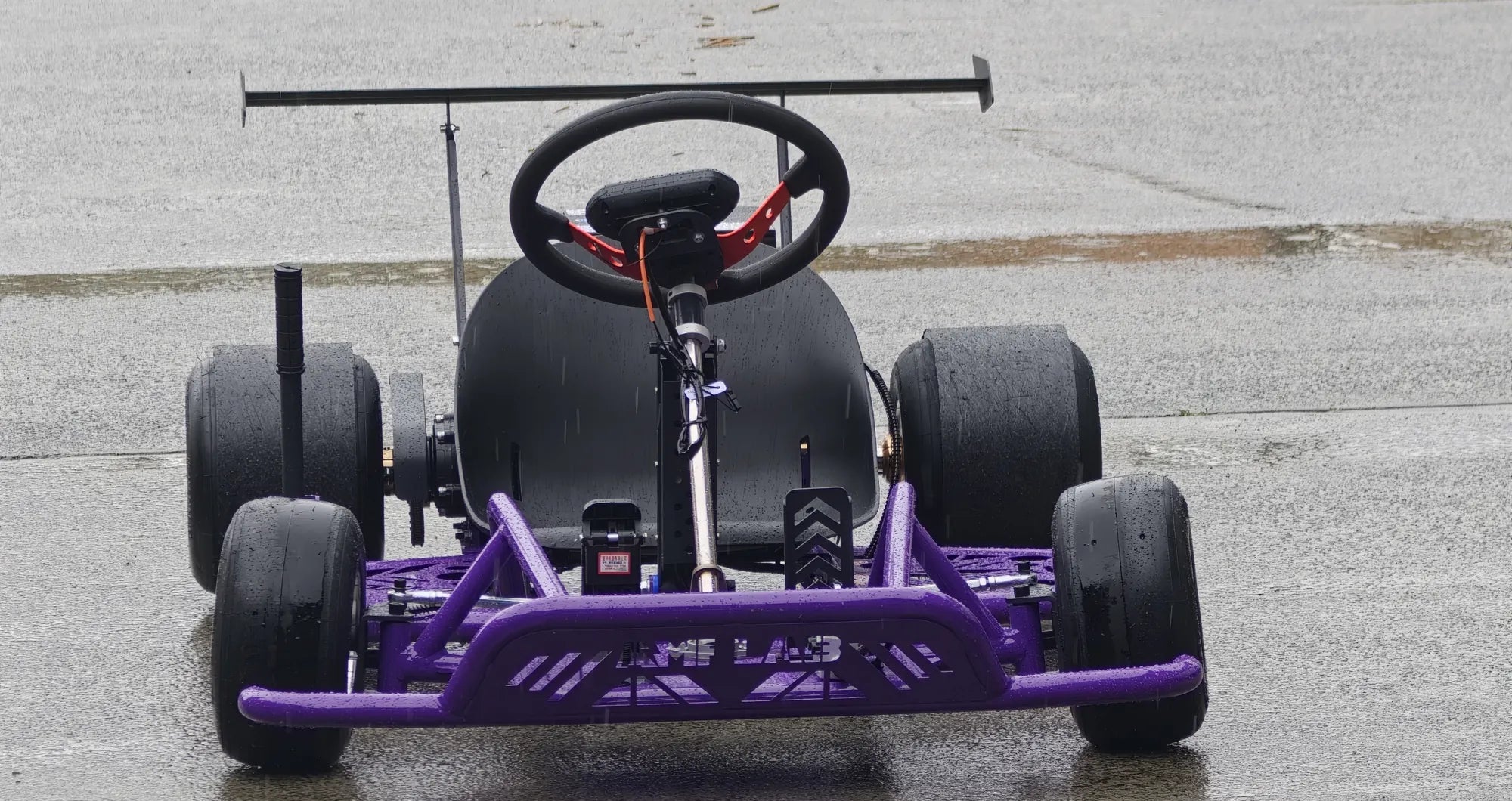 Purple AMPLAB Drift Kart parked on wet concrete, front view showing the AMPLAB bumper, racing wheel and rear wing